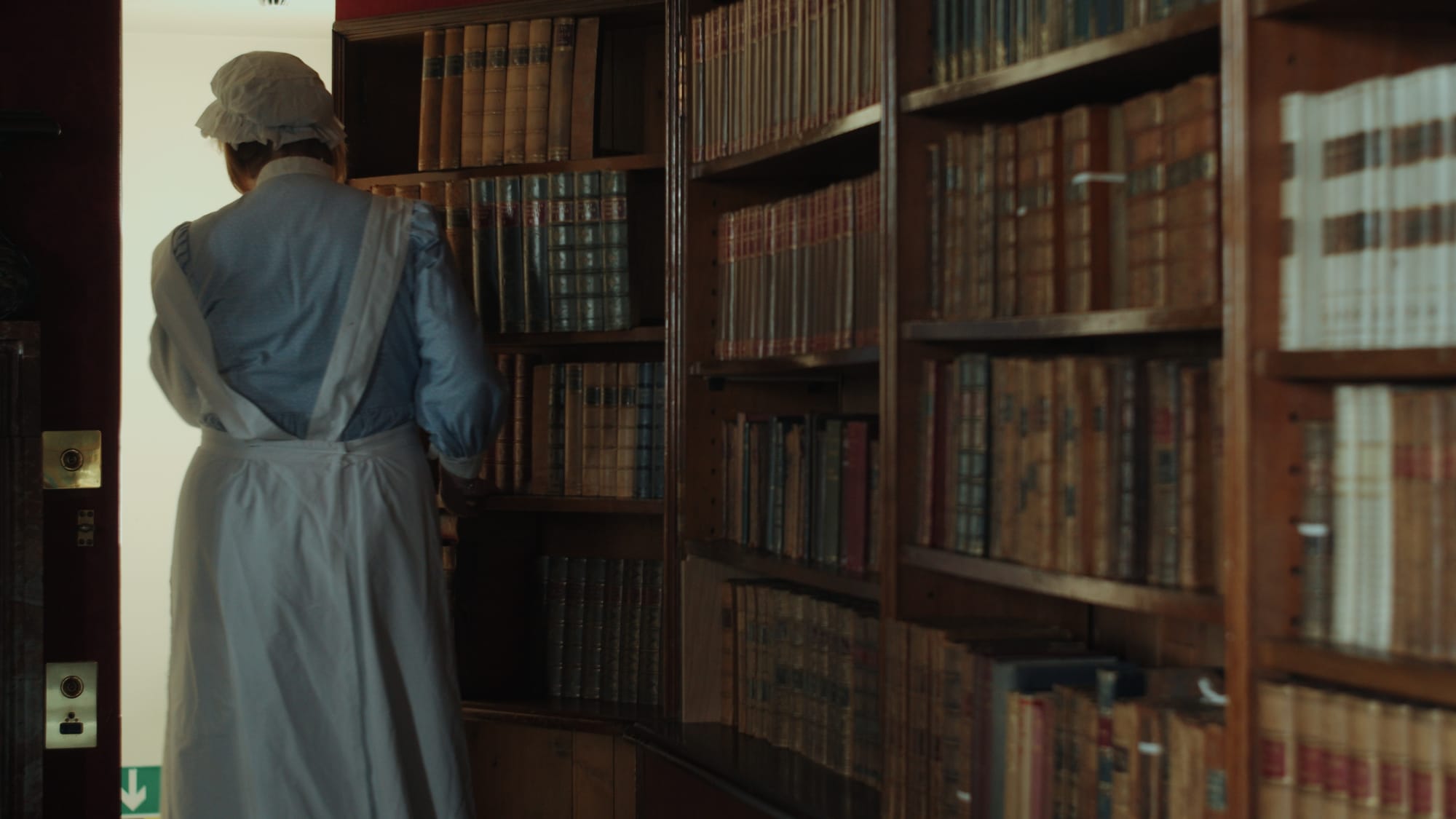 A woman dressed as a maid stands in front of a tall wooden bookshelf filled with old books at Lauriston Castle in Edinburgh